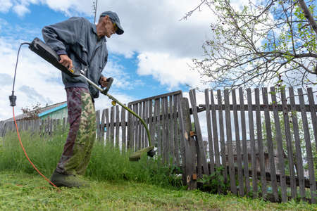 An old man with an electric lawn mower trimmer cuts fresh green grass by the fence wearing glasses and a cap in the village.の写真素材