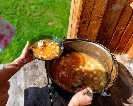 The girl cook outdoors puts soup with meat and vegetables borscht from a large cauldron pan on the wooden floor of the house next to a grass field.の写真素材