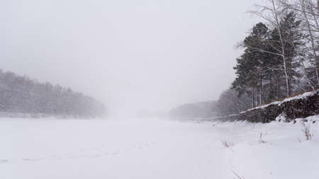A heavy snowstorm covers the tracks on the ice of the river in winter in the middle of two banks with trees and a cliff.の写真素材