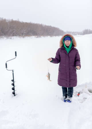 Asian girl is smiling and rejoicing on the ice of the river in the winter holding a fish caught perch on a fishing line next to the ice drill and fishing rod.の写真素材