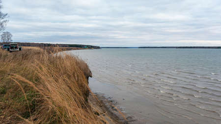 Behind the dry grass, shaking in the wind growing on a cliff above the waves of the troubled dirty sea, there are cars and holiday home recreation on the horizon.の写真素材