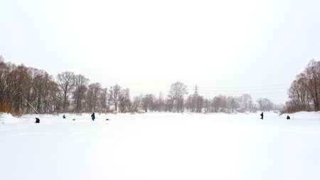 Figures fishermen in the winter in the distance go in search of fish, sit and fish on the ice of the river in a snowstorm against the trees of the forest in Siberia.の写真素材