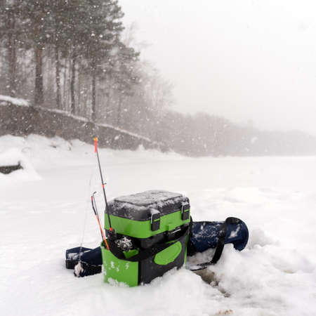 Fishing reel ice fishing rod for fishing spinners sticking out of the angler box in the hole near the cover of the ice under winter Blizzard in the forest.の写真素材
