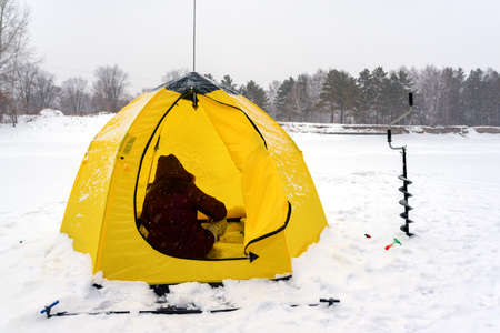 Girl angler winter back in the hood sitting in a tent and ice screws on the background of snow and forest in Russia.の写真素材