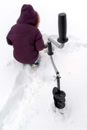 Ice drill for winter fishing with screws stands in front of the girl sitting and fishing holes on the ice of the river in white snow.の写真素材