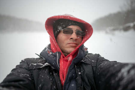 Selfie photo of a man in dark glasses with one eye closed in a hood warmly dressed in winter in a snow storm on the background of a river and forest in the snow.の写真素材