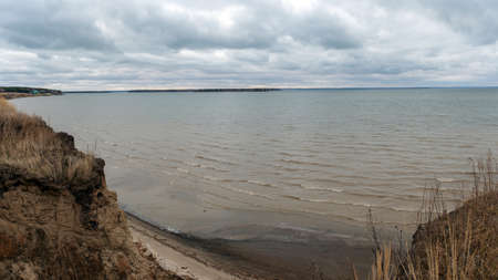 The calm waves of the autumn sea wash the sand on the shore under the cliff with houses on the edge in the distance under the cloudy sky.の写真素材