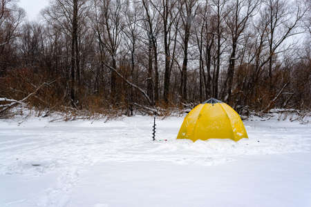 Yellow fishing winter tent stands with an ice-hole in the winter on the ice near the edge of the forest on a snow storm.の写真素材