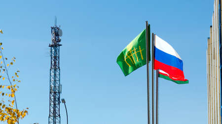 Three flags of Russia, Yakutia, and ulus Suntar are developing in the wind on the background of a radio tower next to the administration building on the background of blue sky and yellow birch.の写真素材
