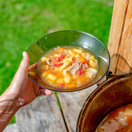 The girl's hands outdoors in the field holding a transparent plate of soup borscht with meat and vegetables on the background of a cauldron with food outdoors in a wooden house.の写真素材