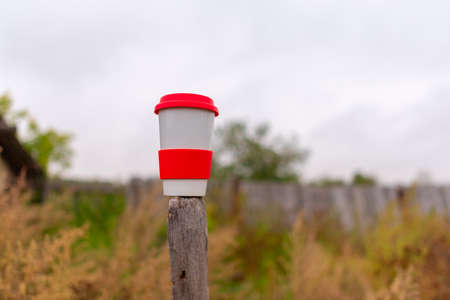 A white ceramic mug with a red lid stands with a drink on a wooden pole in the village in autumn against the background of grass and sky.の写真素材