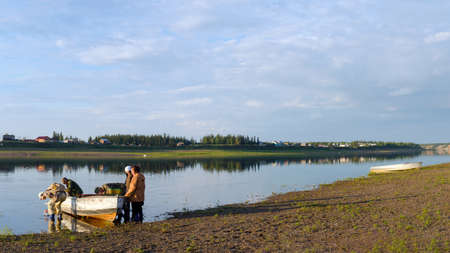 A group of Yakuts sits in a boat standing at the Bank of the river Vilyuy before sailing to the shore with the houses of the village at sunset.の写真素材