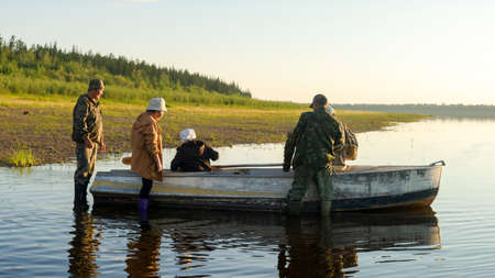 A group of Yakuts stands by a boat in the water of the river Viluy, before sailing to the opposite shore with the houses of the village at sunset.の写真素材
