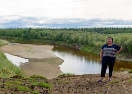Full Yakut Asian girl posing for a photo stands on the edge of a cliff on the background of taiga forest and river in the North of Yakutia.の写真素材