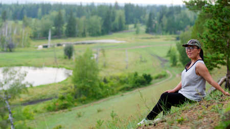 Yakut Asian girl sitting on a hillside smiling on the background of the taiga forest of Yakutia.の写真素材