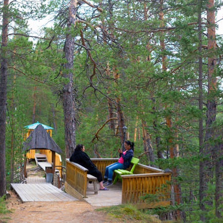Two Yakut girls are resting on a bench in the forest in the Park playing phone in the Northern taiga.の写真素材