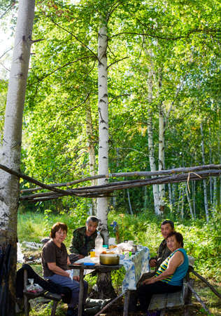 Two pairs of elderly Yakuts sit at a table with food in a place to rest in the wild taiga of the birch forest of the North in the afternoon.の写真素材