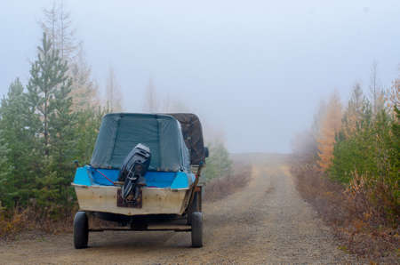 A boat on a trailer with a motor is on the road in the spruce taiga in the morning fog with yellow...