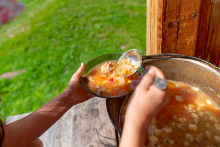 The girl cook outdoors puts soup with meat and vegetables borscht from a large cauldron pan on the wooden floor, in the house next to the grass field, into plates.の写真素材