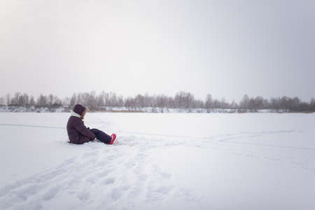 A lonely girl sits on the snow ice of a winter lake, clasping her knees with her hands.