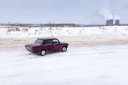 Russia, Novosibirsk - November 30, 2019. Russian old crimson car "VAZ-Zhiguli" goes drifting, raising snow Blizzard on the turn for the pipes of the thermal power plant.のeditorial素材