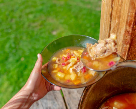 Hands girl cooks outdoors puts soup with meat and vegetables and potatoes, beet borscht from a large cauldron pan on the wooden floor, in a house next to a grass field in the woods, in transparent plates.の写真素材