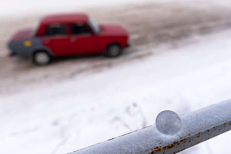 A small glass ball standing calmly on a railing in the snow reflects a silhouette with a fast white drift racing car on the road in winter.の写真素材
