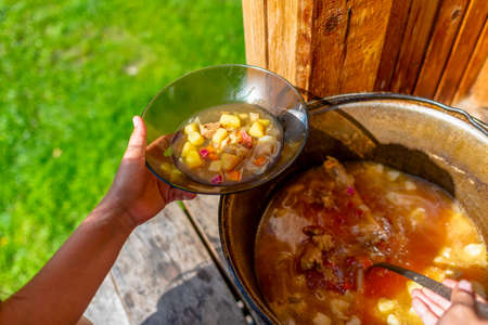 The hands of a girl cook outdoors lays soup with meat and vegetables and potato borscht from a large cauldron pan on the wooden floor, in a house next to a grass field in the woods, in transparent plates.の写真素材