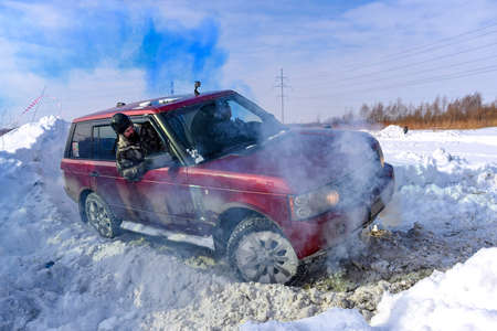 Russia, Novosibirsk-March 13, 2020. A "Range Rover 4x4" SUV is actively driving on a snowy mountain in colored blue smoke, and a man outside in a hard hat is watching the road.のeditorial素材