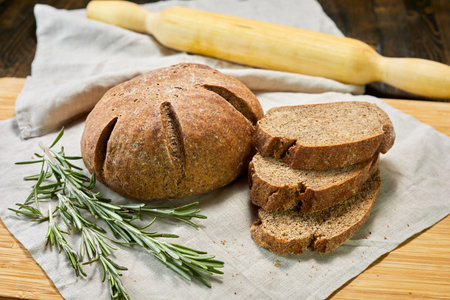 Freshly baked rustic rye bread with rosemary on linen cloth. Sliced loaf reveals dense, grainy...