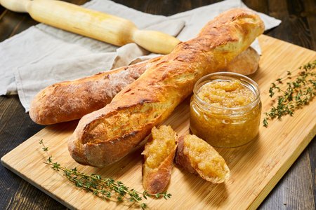 Fresh baguettes with apple or quince jam on wooden board. Sliced bread spread with sweet preserve, garnished with thyme. Rolling pin and linen napkin in background. Perfect for breakfast, brunch or bakery content.の写真素材