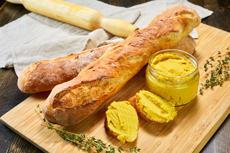 Two golden, crusty French baguettes arranged on a wooden board, one sliced and generously spread with creamy yellow mustard. A small glass jar of the same mustard sits beside themの写真素材