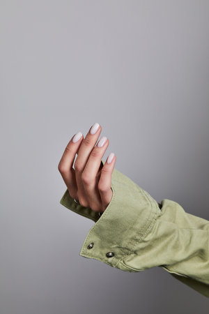 Womanâs hand with soft white manicure gripping the cuff of a light green denim jacket against a neutral grey backgroundの写真素材