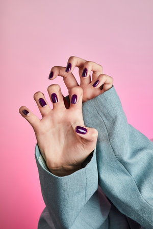 Elegant hands with deep purple manicure peeking from a light blue blazer sleeve against a vibrant pink backgroundの写真素材