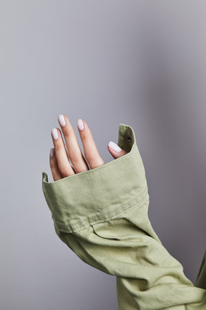 Womanâs hand with soft white manicure gripping the cuff of a light green denim jacket against a neutral grey backgroundの写真素材