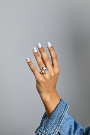 Womanâs hand with light blue manicure and a sculptural silver ring, posed against a neutral grey background with denim sleeveの写真素材