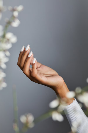 Elegant hand with pristine white manicure and soft sweater sleeve against a gray backdrop with floral bokehの写真素材