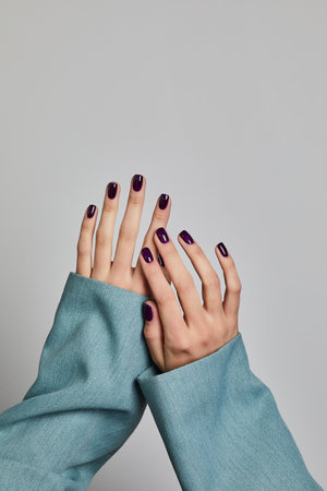 Two elegant hands with glossy purple manicure and light blue sleeves posed against a clean gray backgroundの写真素材