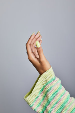 Stylish female hand with lime-green manicure and silver ring emerging from a pastel striped knit sleeve on gray backgroundの写真素材