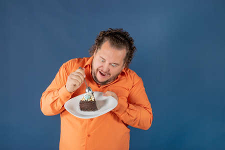 Funny fat man in orange shirt with a piece of chocolate cake on a plate. Birthday celebrationの写真素材