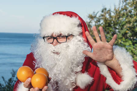 Fat man in glasses dressed as Santa holds tangerines on the oceanの写真素材