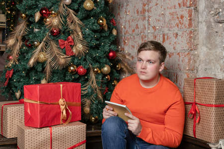 Handsome guy is writing a letter to Santa sitting under the tree surrounded by boxes of giftsの写真素材