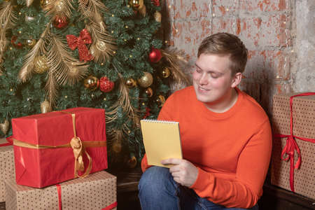 Handsome guy is writing a letter to Santa sitting under the tree surrounded by boxes of giftsの写真素材