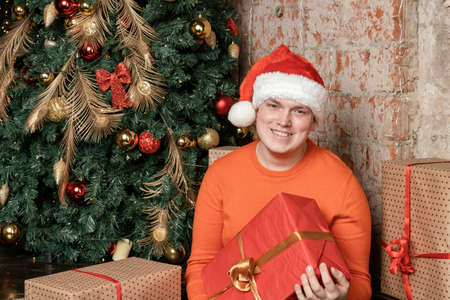 Handsome guy in santa claus hat holding box with gift sitting under the tree surrounded by boxes of giftsの写真素材
