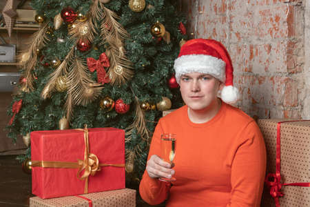 Handsome guy in santa claus hat holds a glass of champagne sitting under the tree surrounded by boxes of giftsの写真素材