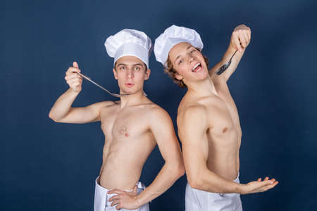 Two handsome sexy chefs with apron on naked muscular body with kitchen utensils on blue backgroundの写真素材
