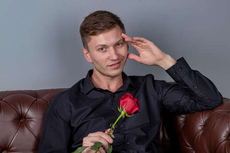 Handsome man wearing black shirt sitting on the leather sofa holding a roseの写真素材