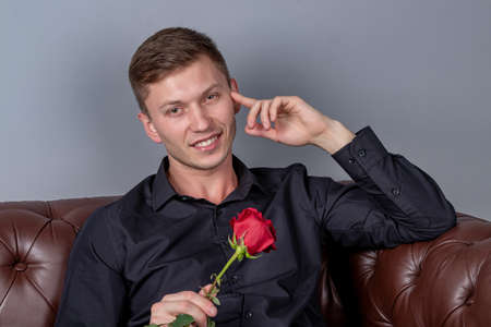 Handsome man wearing black shirt sitting on the leather sofa holding a roseの写真素材