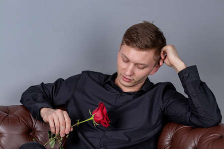 Handsome man wearing black shirt sitting on the leather sofa holding a roseの写真素材