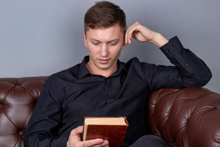 Young man is sitting on a sofa and reading a bookの写真素材
